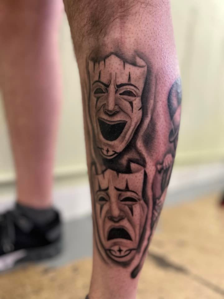 A man has a tattoo of two comedy masks on his leg.