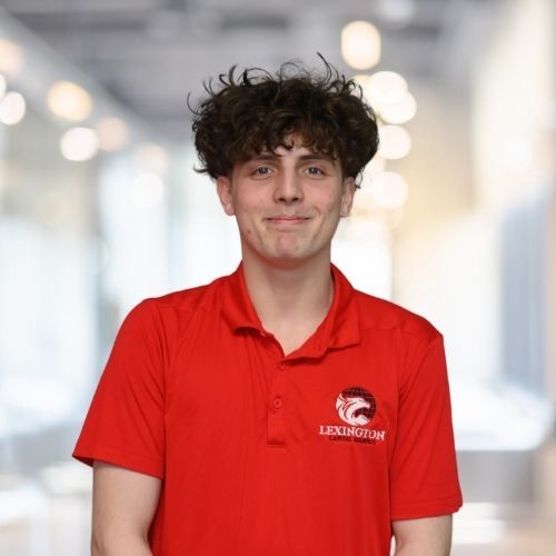 Woman in red polo shirt smiles at the camera, wearing a Lexington logo. Blurred indoor background.