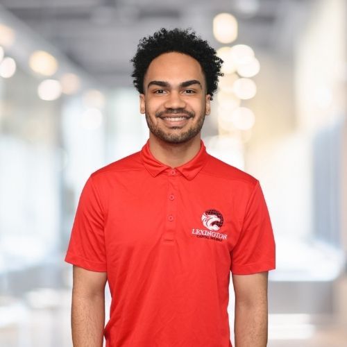 Woman in red polo shirt smiles at the camera, wearing a Lexington logo. Blurred indoor background.