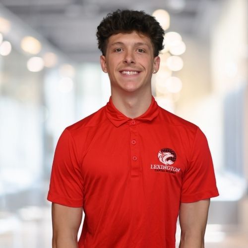 Woman in red polo shirt smiles at the camera, wearing a Lexington logo. Blurred indoor background.