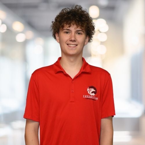 Woman in red polo shirt smiles at the camera, wearing a Lexington logo. Blurred indoor background.