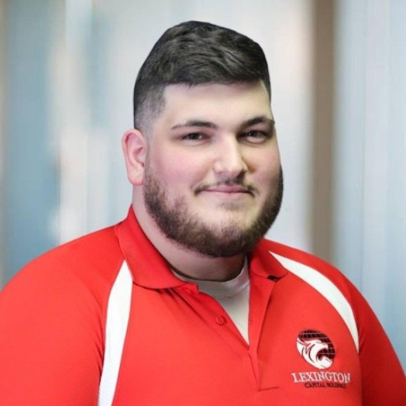 Woman in red polo shirt smiles at the camera, wearing a Lexington logo. Blurred indoor background.