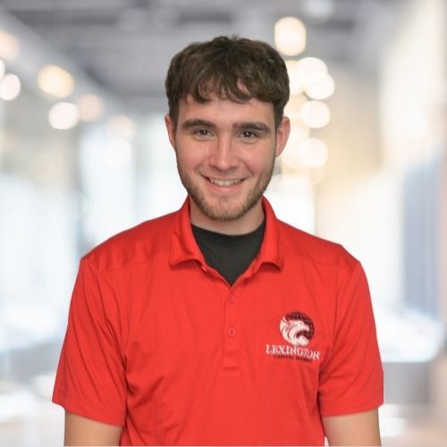 Woman in red polo shirt smiles at the camera, wearing a Lexington logo. Blurred indoor background.
