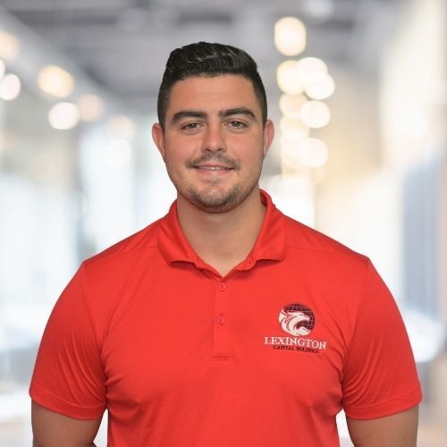 Woman in red polo shirt smiles at the camera, wearing a Lexington logo. Blurred indoor background.