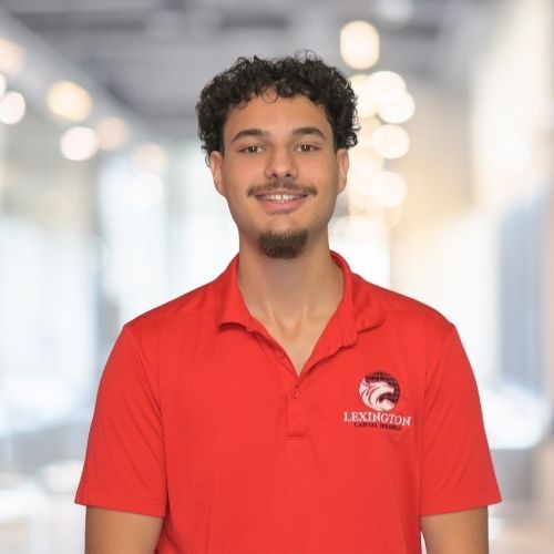 Woman in red polo shirt smiles at the camera, wearing a Lexington logo. Blurred indoor background.