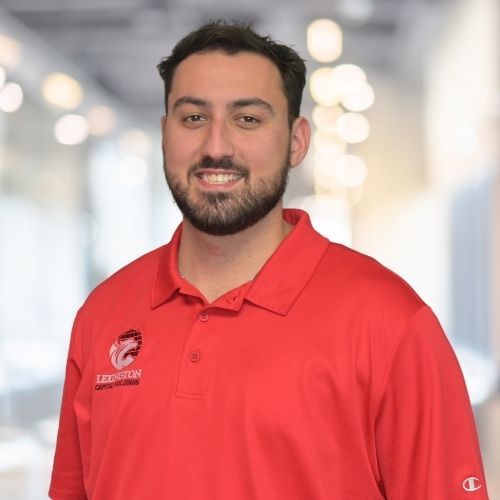 Woman in red polo shirt smiles at the camera, wearing a Lexington logo. Blurred indoor background.
