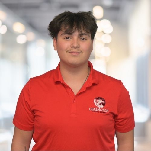 Woman in red polo shirt smiles at the camera, wearing a Lexington logo. Blurred indoor background.