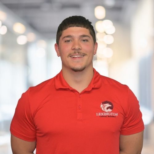 Woman in red polo shirt smiles at the camera, wearing a Lexington logo. Blurred indoor background.