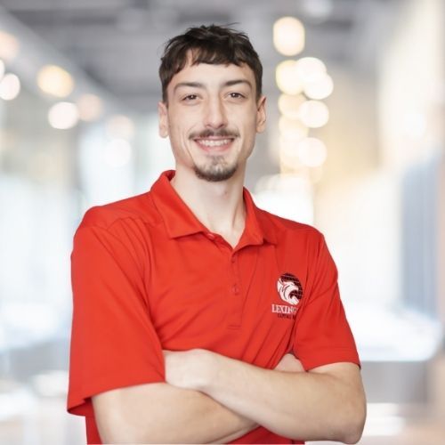 Woman in red polo shirt smiles at the camera, wearing a Lexington logo. Blurred indoor background.