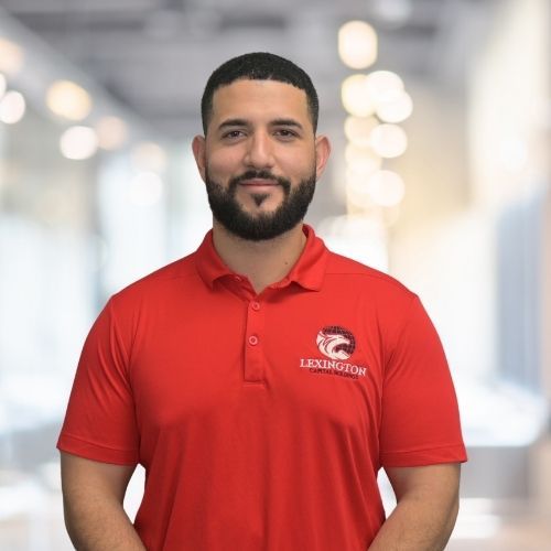 Woman in red polo shirt smiles at the camera, wearing a Lexington logo. Blurred indoor background.