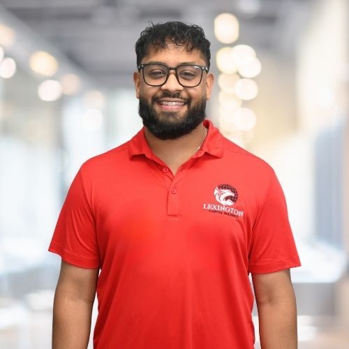 Woman in red polo shirt smiles at the camera, wearing a Lexington logo. Blurred indoor background.