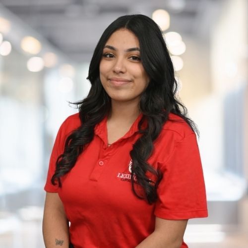 Woman in red polo shirt smiles at the camera, wearing a Lexington logo. Blurred indoor background.