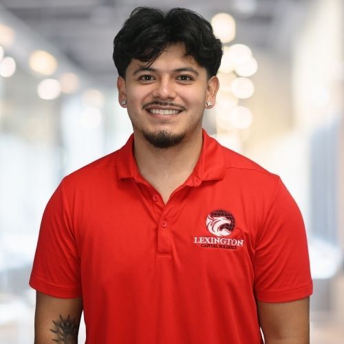 Woman in red polo shirt smiles at the camera, wearing a Lexington logo. Blurred indoor background.