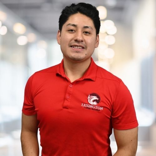 Woman in red polo shirt smiles at the camera, wearing a Lexington logo. Blurred indoor background.