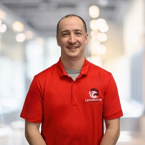 Woman in red polo shirt smiles at the camera, wearing a Lexington logo. Blurred indoor background.