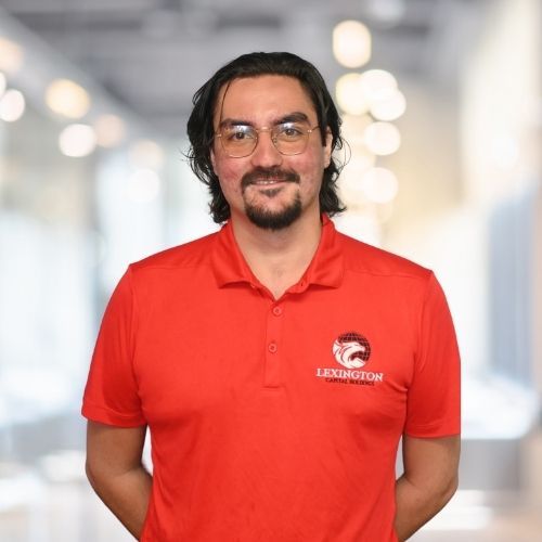 Woman in red polo shirt smiles at the camera, wearing a Lexington logo. Blurred indoor background.