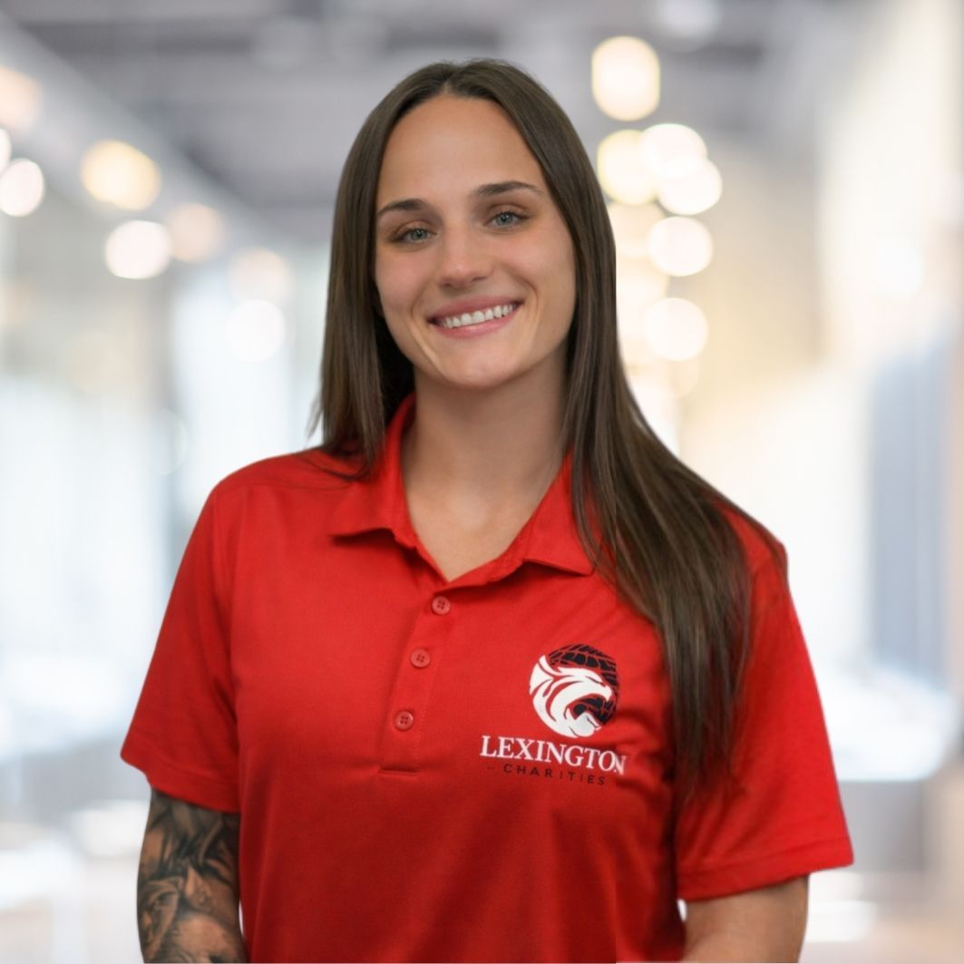 Woman in red polo shirt smiles at the camera, wearing a Lexington logo. Blurred indoor background.