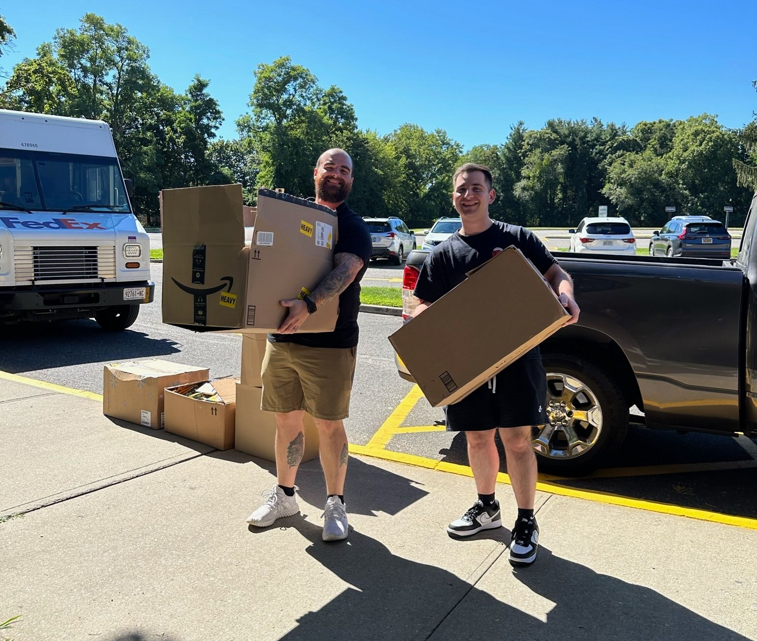 Two people holding Amazon boxes next to a delivery truck and a pickup truck. Sunny day.