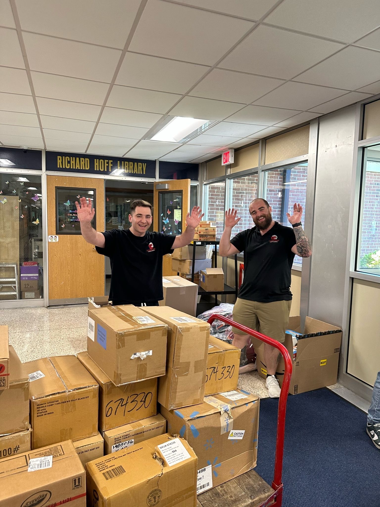 Two men wave, standing by a stack of cardboard boxes in a hallway.
