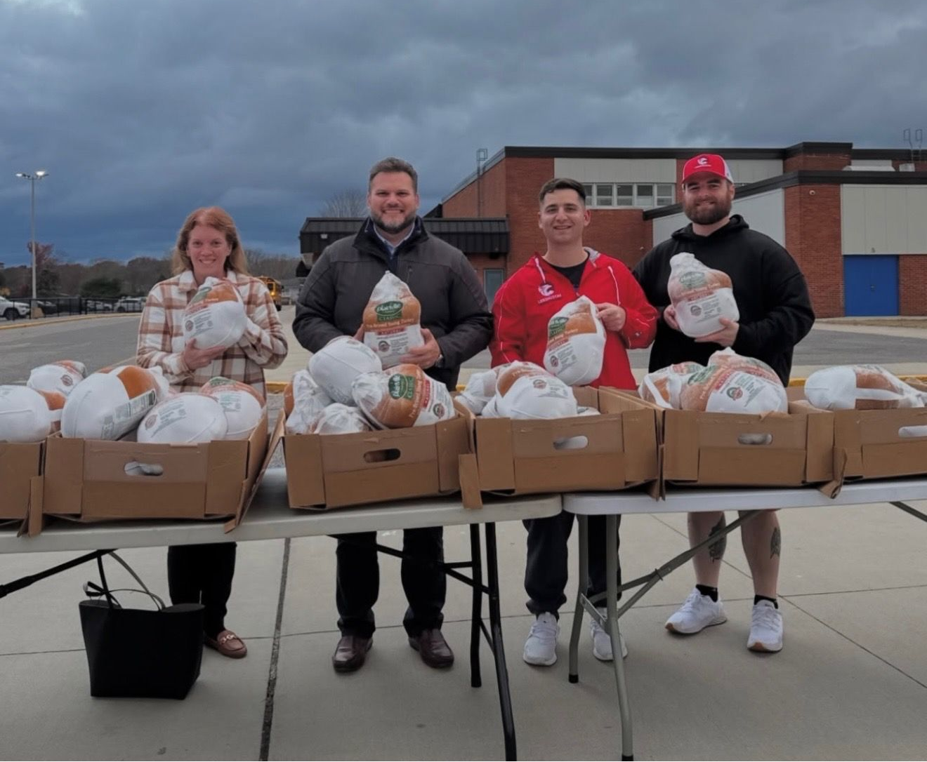 Four people standing behind tables laden with turkeys, likely for a food drive, outside a building.