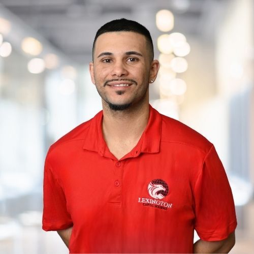 Woman in red polo shirt smiles at the camera, wearing a Lexington logo. Blurred indoor background.