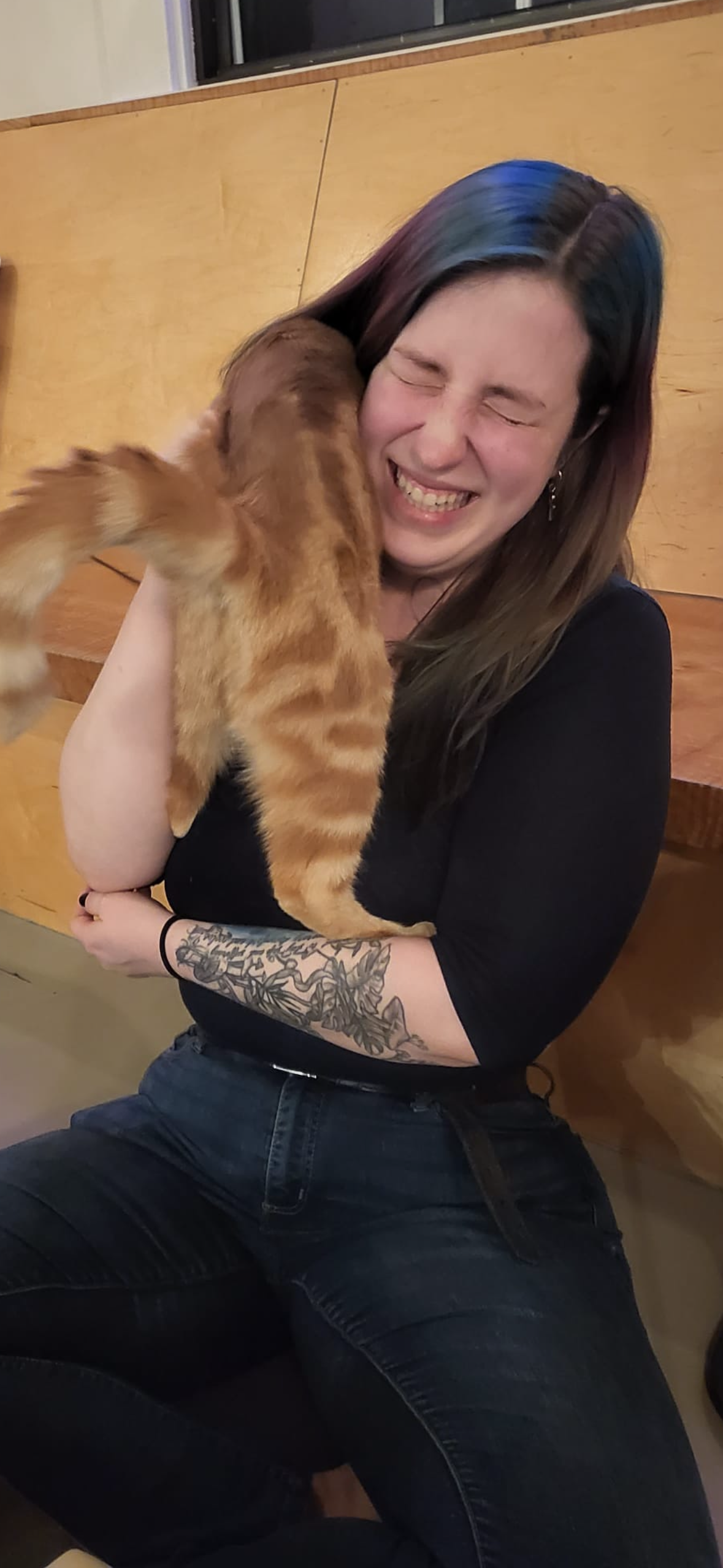 Woman laughing, holding a ginger cat. The cat's leg is near the woman's face. They are indoors.