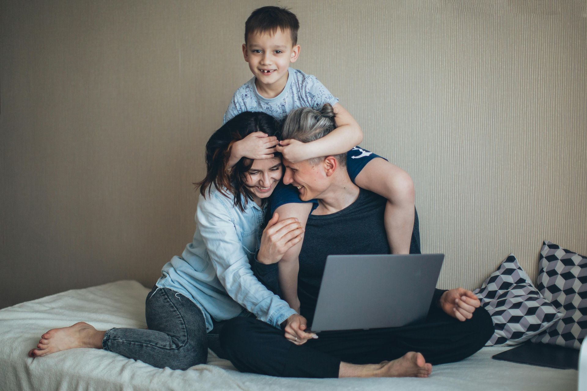 Family on a bed, looking at laptop. Boy on father's shoulders, mother touching father's head, smiling.