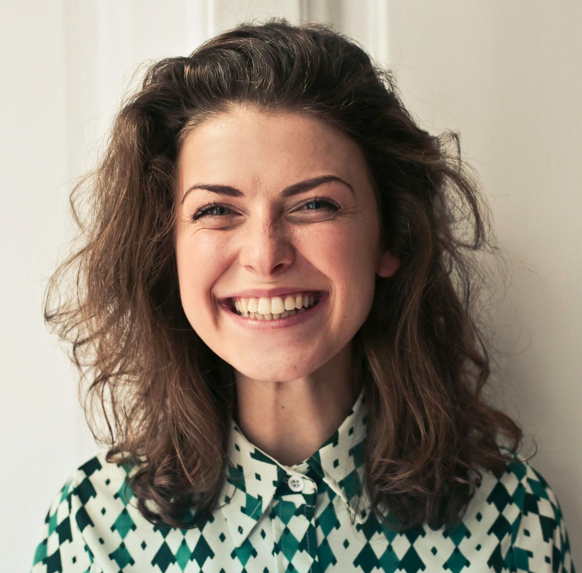Woman with curly brown hair, wearing a patterned green shirt, smiling widely.