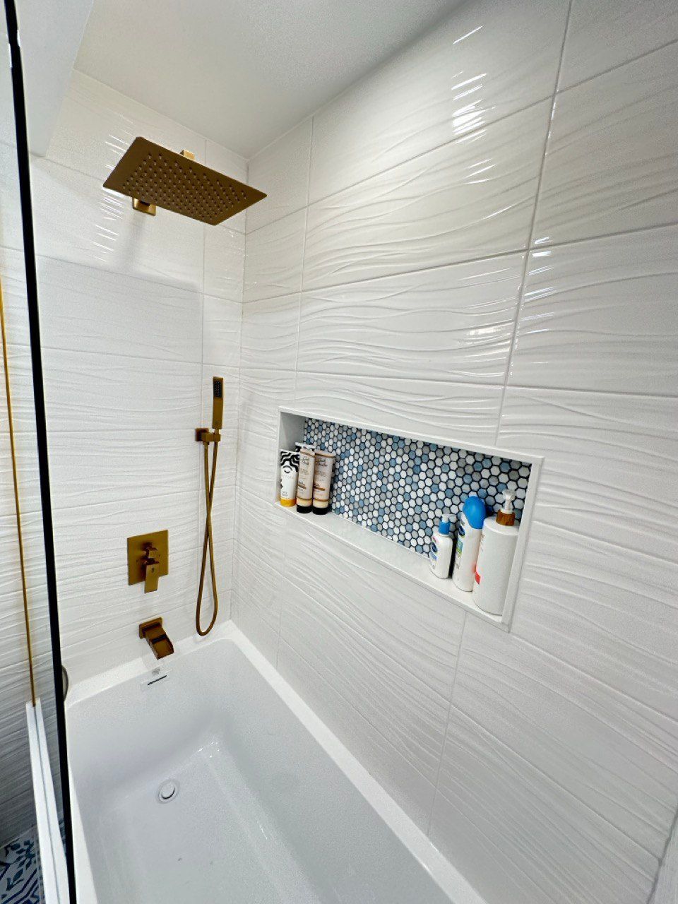 Hallway bathroom remodeling in Sykesville featuring white wavy wall tile with a large horizontal niche accented in blue tile, deep soaking tub with brushed gold faucet and handheld, rain shower head, fixed glass panel, blue floor tile design, custom vanity with quartz top, and a quiet exhaust fan.