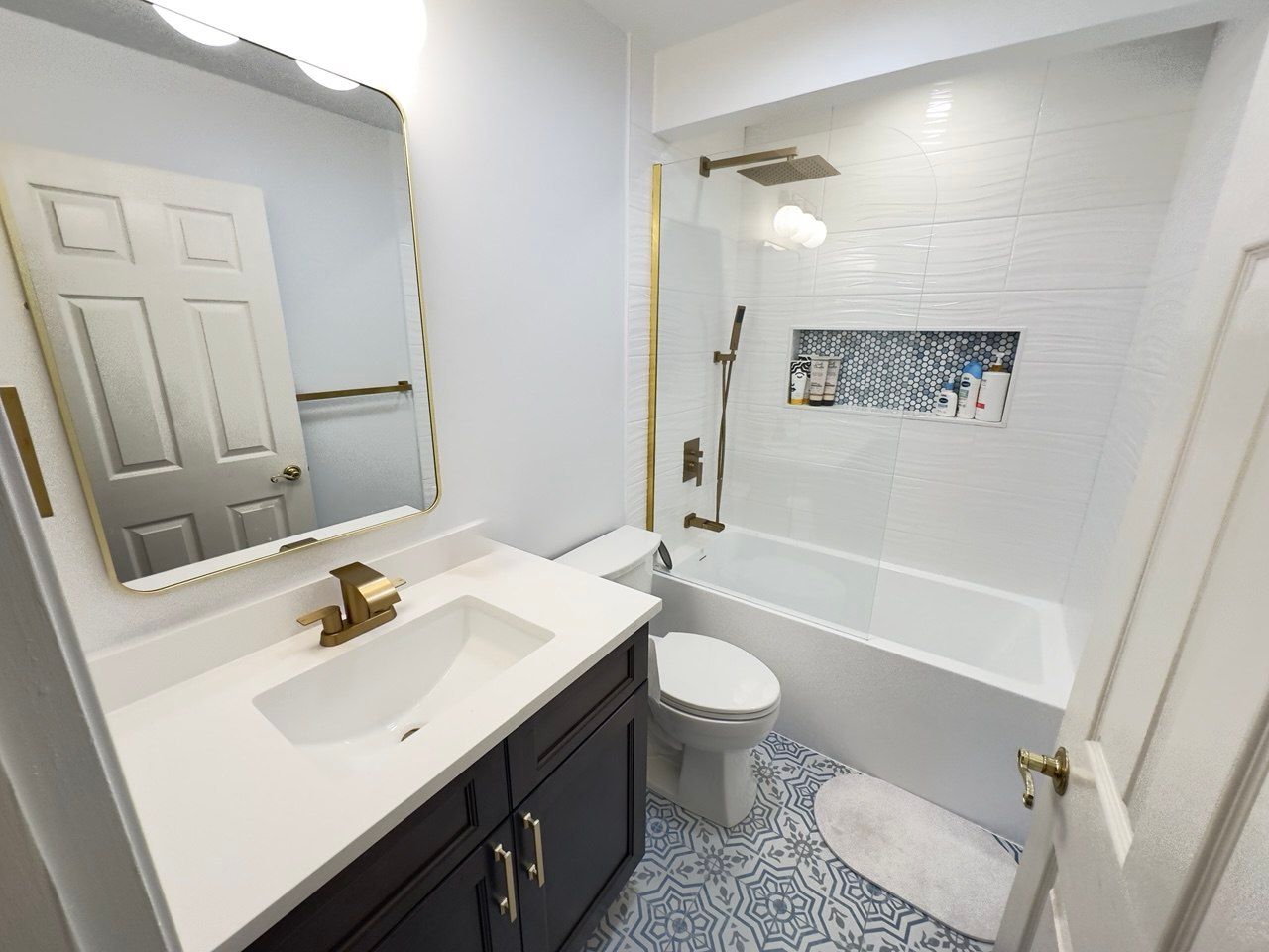 Hallway bathroom remodeling in Sykesville featuring white wavy wall tile with a large horizontal niche accented in blue tile, deep soaking tub with brushed gold faucet and handheld, rain shower head, fixed glass panel, blue floor tile design, custom vanity with quartz top, and a quiet exhaust fan.