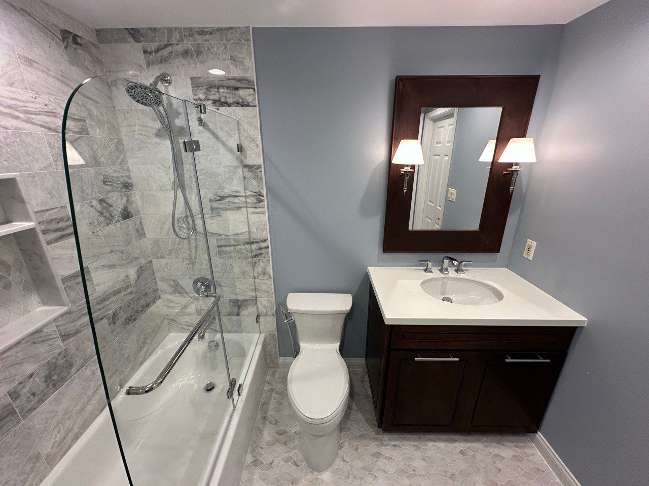 This elegant basement bathroom remodel in Cockeysville features beautiful marble wall tile with a built-in niche, adding both function and luxury. The shower includes a frameless swing glass door that creates a clean, modern look. A dark vanity contrasts perfectly with the light marble tile, complemented by stylish wall sconces on each side of the mirror. The marble tile flooring ties the whole space together, creating a timeless and sophisticated design.