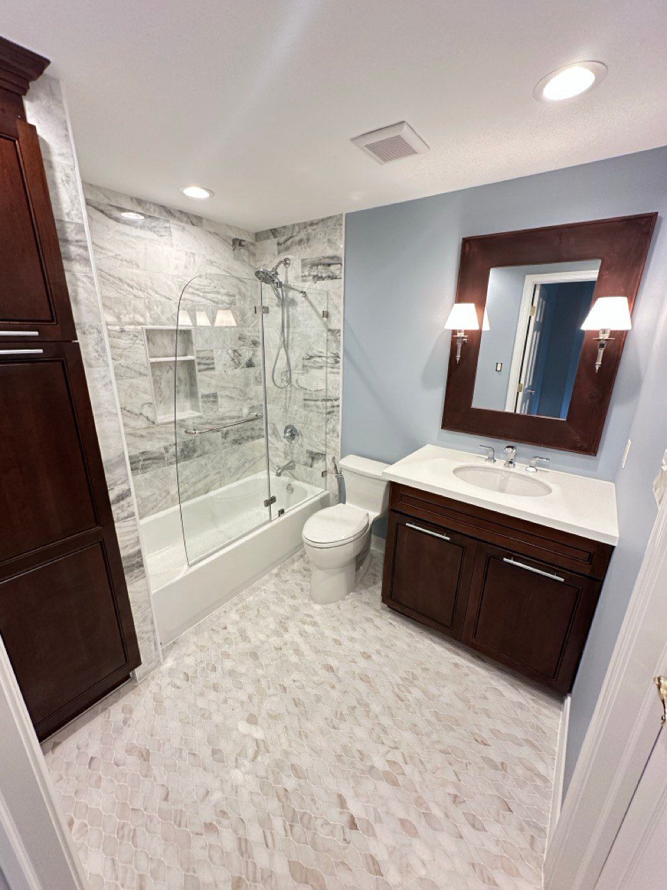 This elegant basement bathroom remodel in Cockeysville features beautiful marble wall tile with a built-in niche, adding both function and luxury. The shower includes a frameless swing glass door that creates a clean, modern look. A dark vanity contrasts perfectly with the light marble tile, complemented by stylish wall sconces on each side of the mirror. The marble tile flooring ties the whole space together, creating a timeless and sophisticated design.