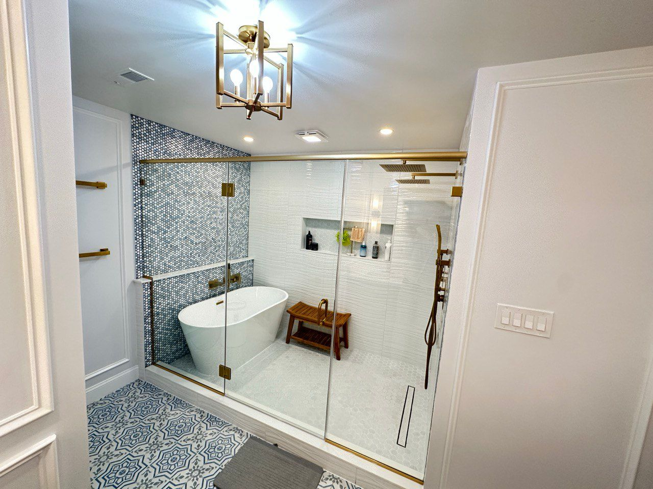 Luxury master bathroom remodeling in Sykesville featuring a spacious 5x10 ft walk-in shower with a freestanding tub inside, dual shower faucets, frameless glass door, and chandelier lighting. Custom blue accent wall tile, blue floor tile, wave-pattern white wall tile, brushed gold fixtures, and large niche complete the elegant design.