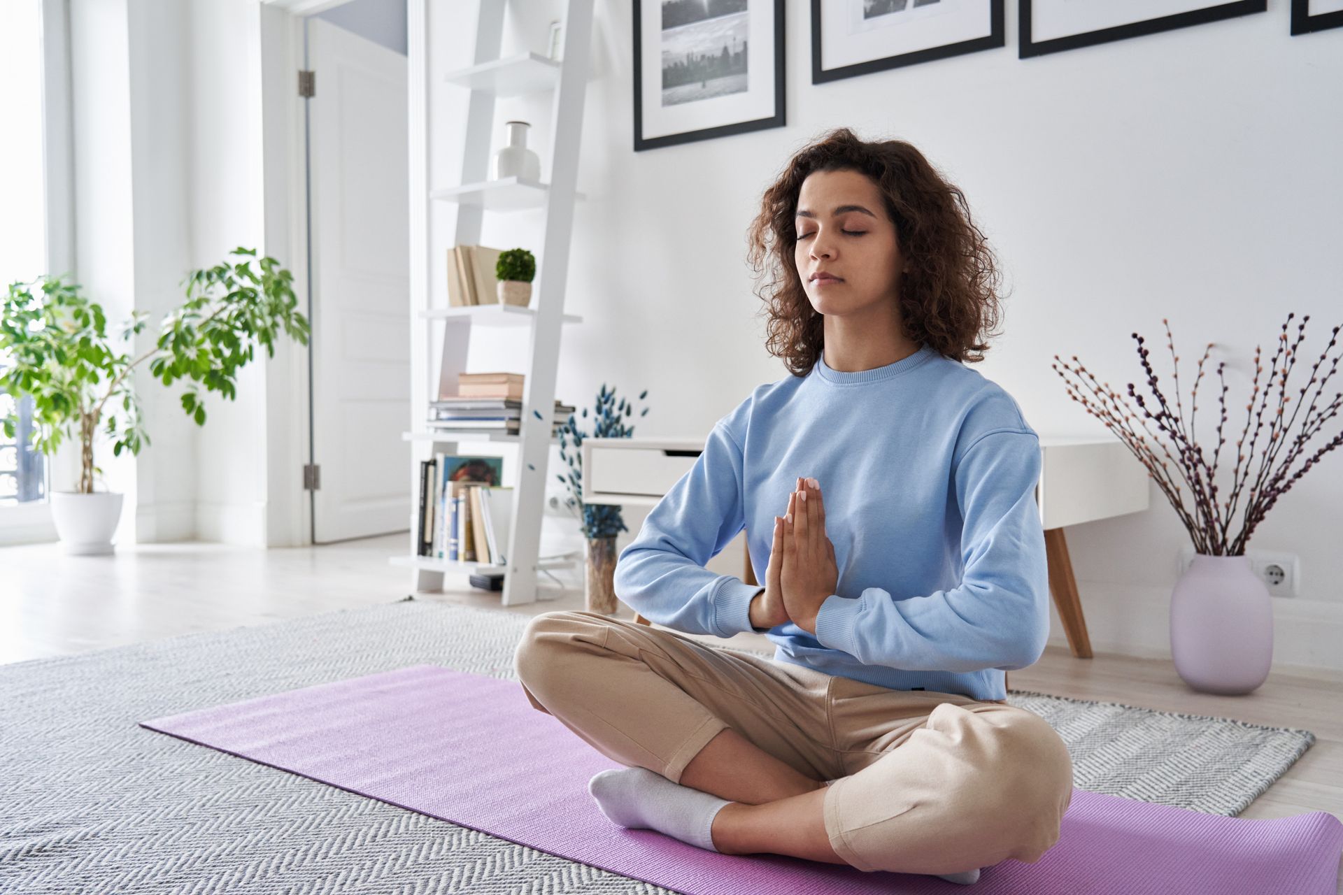 Woman meditating indoors with hands together; sitting on a purple mat.