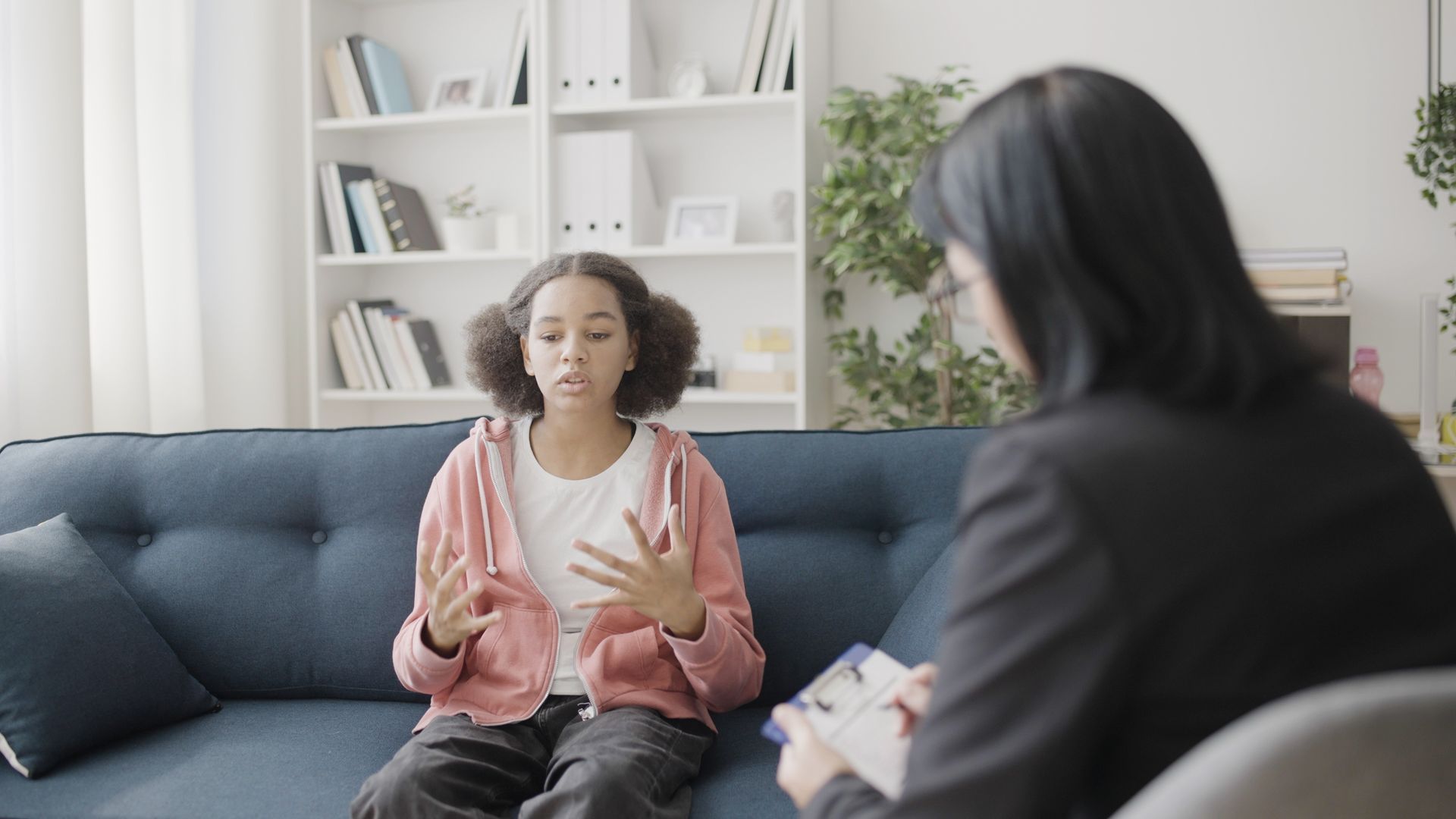 A girl speaks to a female therapist while sitting on a sofa, inside a room.
