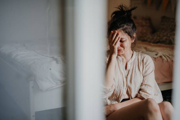 A woman is sitting on a bed covering her face with her hand.
