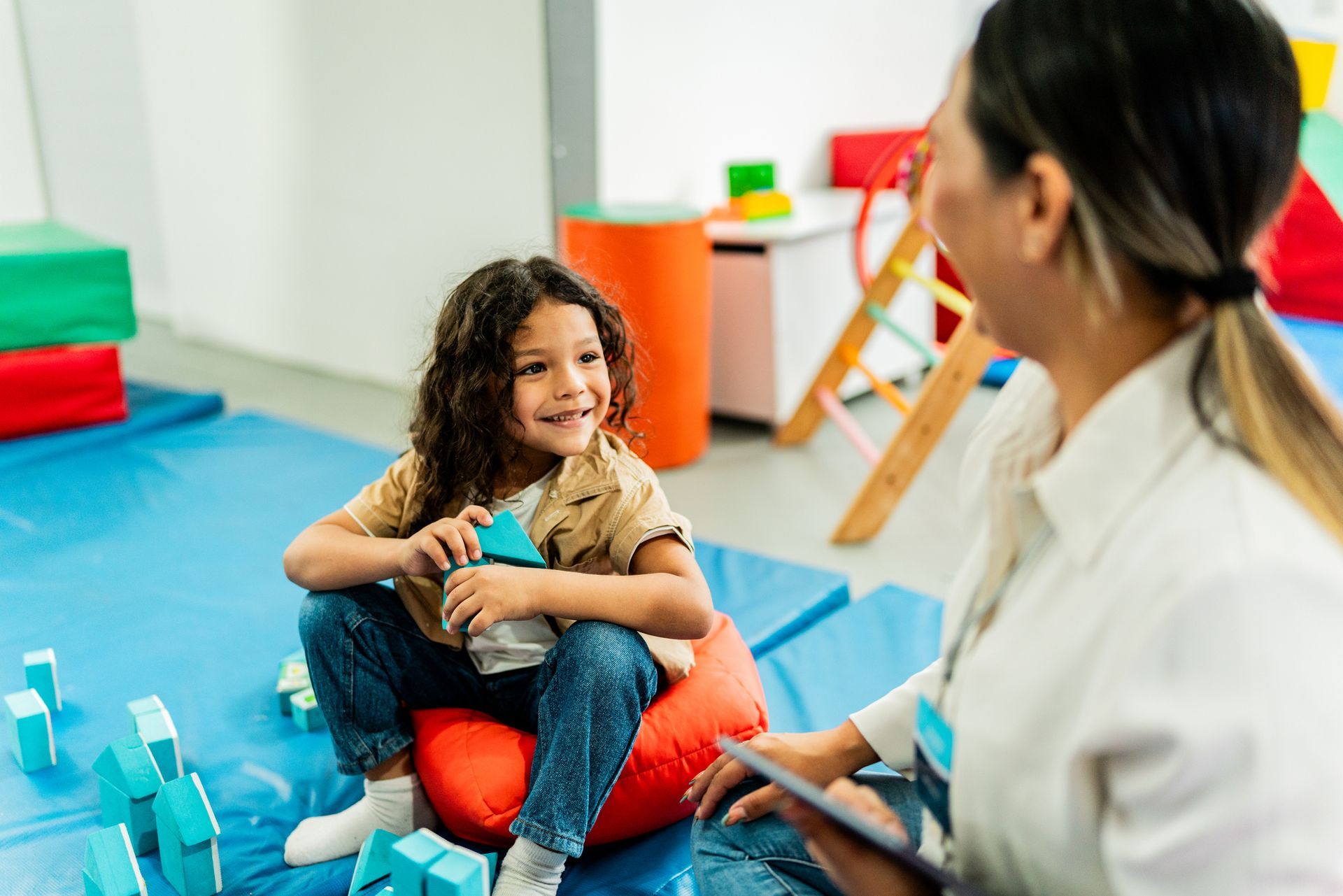 A happy child sits on a cushion as he plays with blocks and talks to a child therapist.
