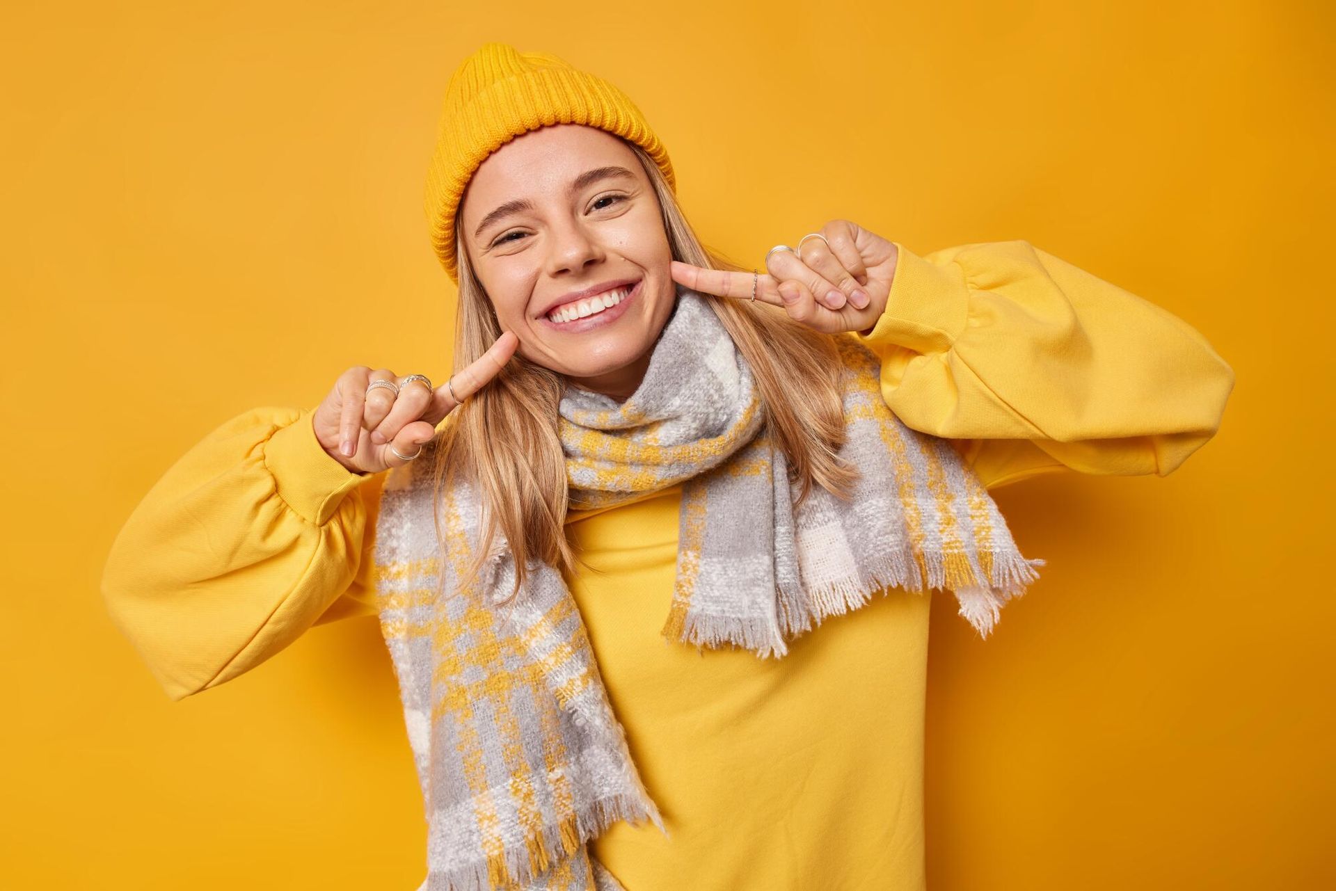 A woman wearing a yellow hat and scarf is smiling and covering her mouth with her hands.