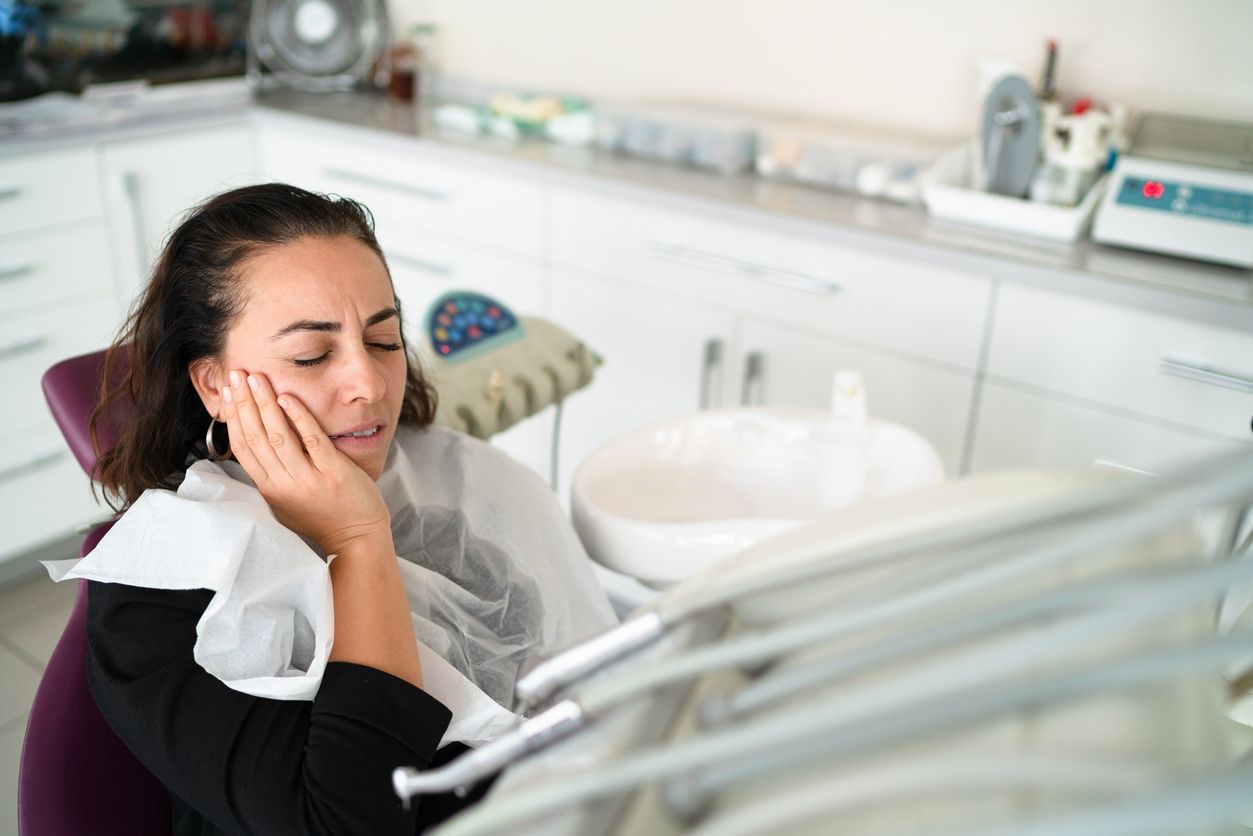 Woman in dentist chair holding cheek, appearing in pain, white and silver dental equipment.