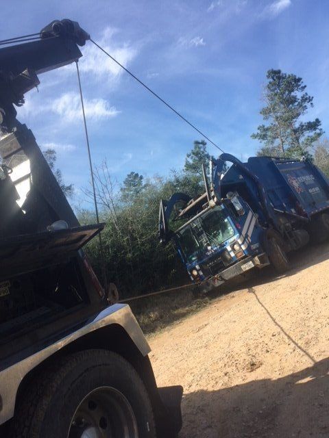 A tow truck pulling a blue and white garbage truck out of a ditch on a dirt road.