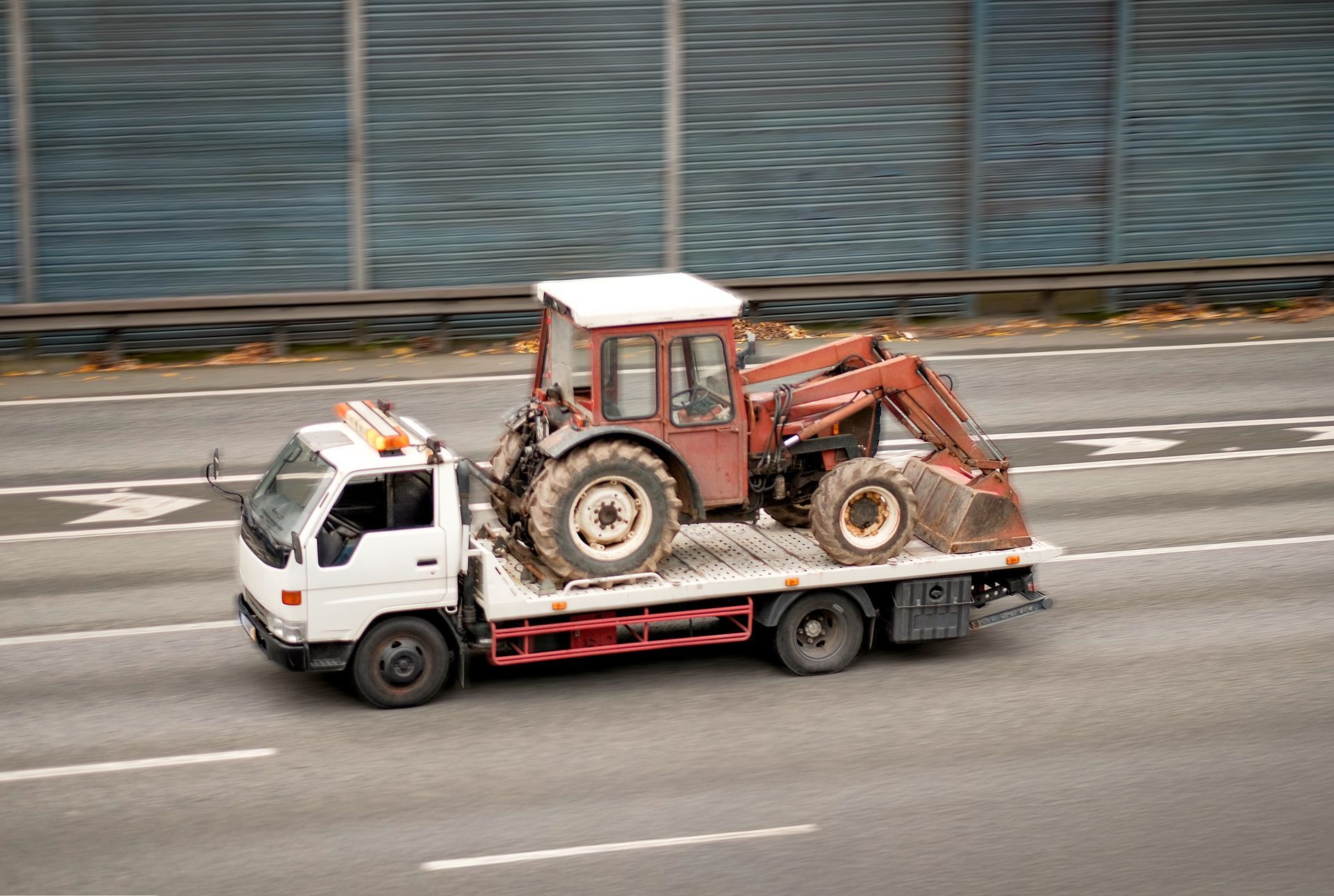 A white tow truck hauling a red tractor by a highway sound wall.