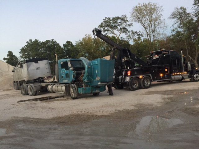 Tow truck hooking up to a disabled semi-truck with a dump trailer in a gravel lot.