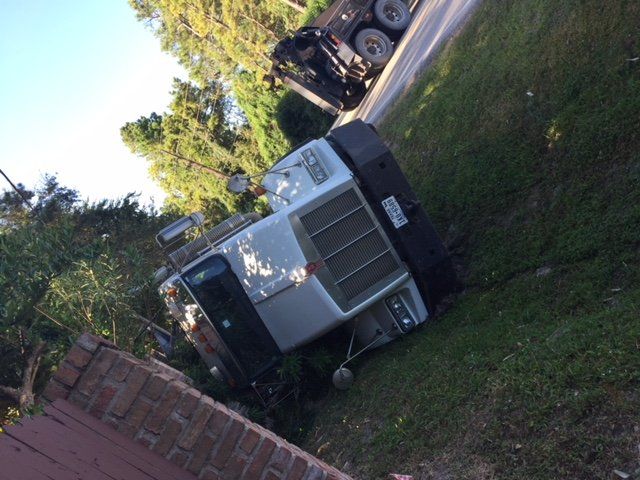 Semi-truck on its side in a ditch next to a road and a brick wall.