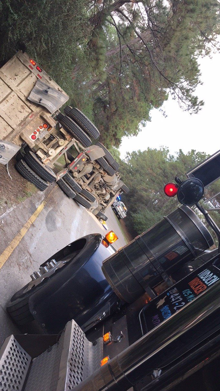 Overturned semi-truck on a wet road, next to a car; trees and a traffic light visible.