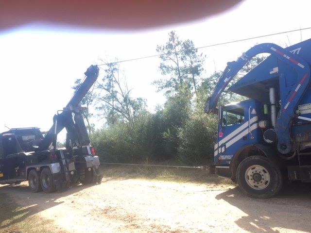 Tow truck preparing to lift a blue and white garbage truck, on a dirt road, trees in the background.