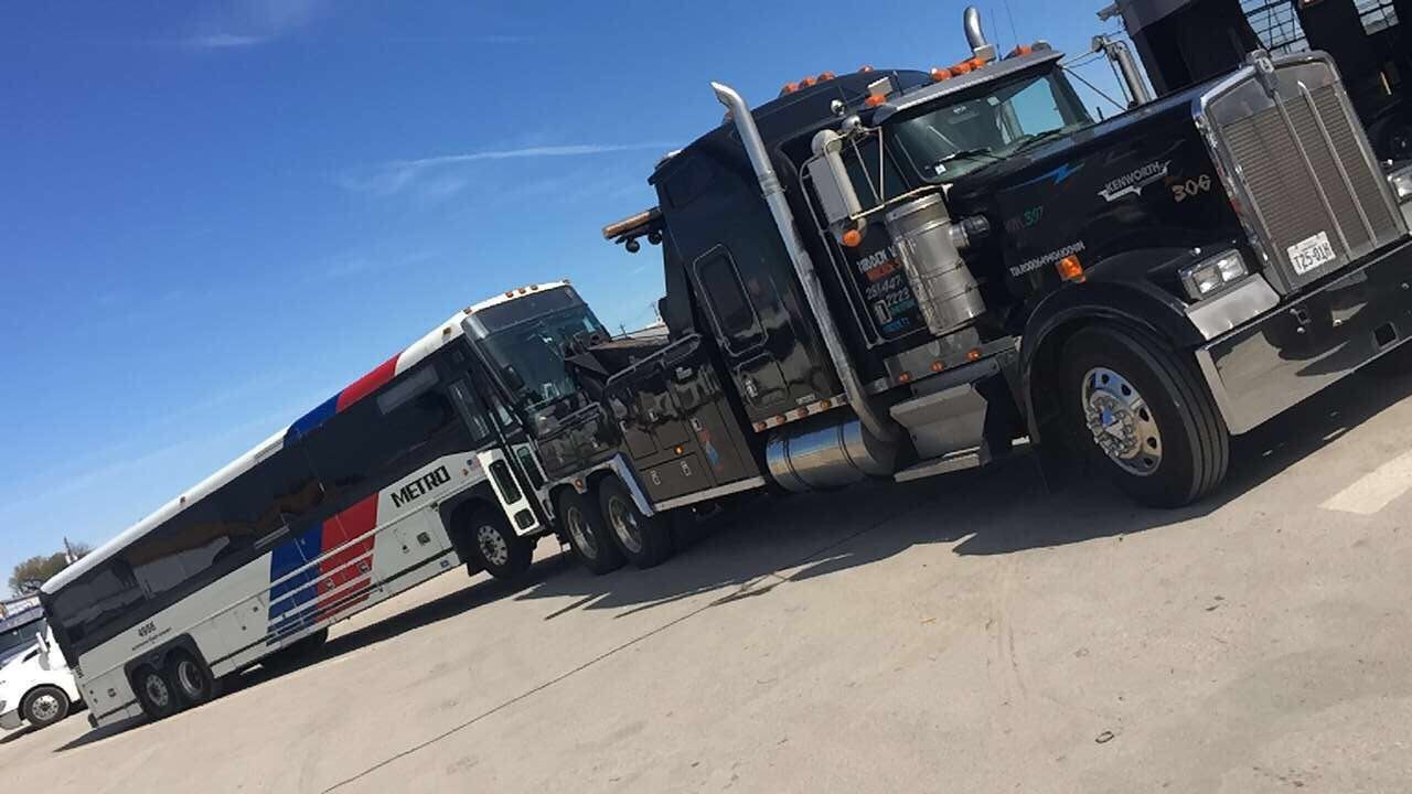 A black tow truck towing a white bus with red and blue stripes, on a clear, sunny day.