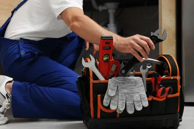 Plumber reaching into toolbox with tools, kneeling under a sink, wearing blue overalls.