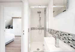 Bright white bathroom with glass shower, sink, and view into bedroom.