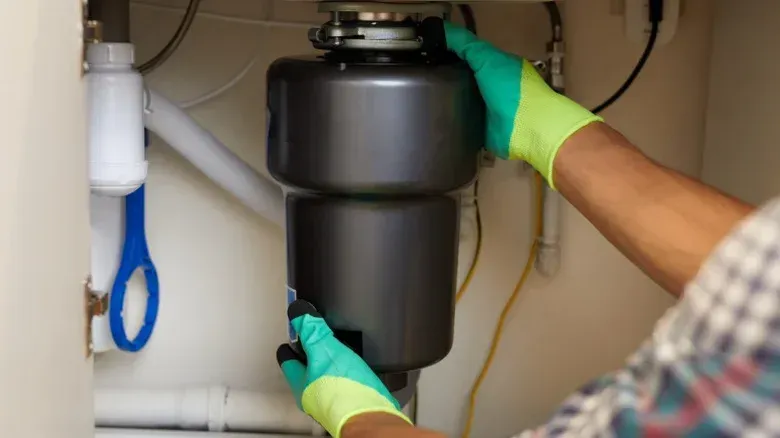 Hands wearing green gloves installing a dark gray garbage disposal under a sink.