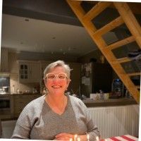 Une femme à lunettes sourit dans une cuisine chaleureuse, éclairée par une lumière tamisée ; une échelle en bois est visible.